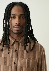 Brunternetternet knap-shirt lavet af blødt stof, med en brystlomme, sorte knapper og forskellige teksturerede mønstre i nuancer af brunt.