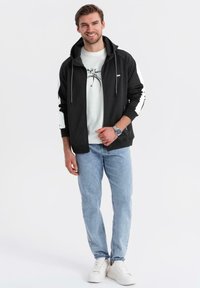 Czarna bluza z zamkiem błyskawicznym i białymi akcentami, noszona na jasnoszarym T-shircie z grafiką. Jasnoniebieskie dżinsy i białe sneakersy dopełniają outfit.