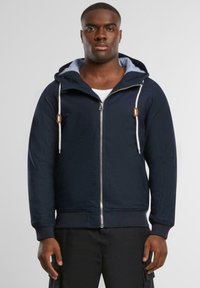 Marineblå hettegenser med glidelås, lett stoff, lommer foran og kontrasterende innersøm. Har en tettsittende design.