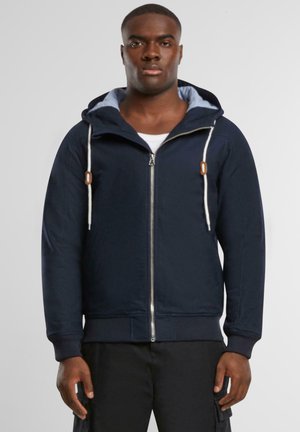 Marineblå hettegenser med glidelås, lett stoff, lommer foran og kontrasterende innersøm. Har en tettsittende design.