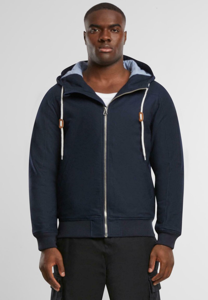 Felpa con zip di colore navy, dotata di cappuccio regolabile, tessuto leggero, tasche frontali e fodera interna a contrasto. Presenta un design aderente.