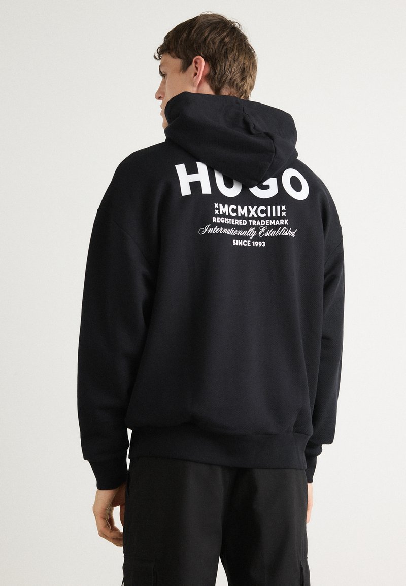 Svart hettegenser laget av mykt stoff, med en stor hvit "HUGO"-logo på baksiden, samt ekstra tekst og snor i hetta.