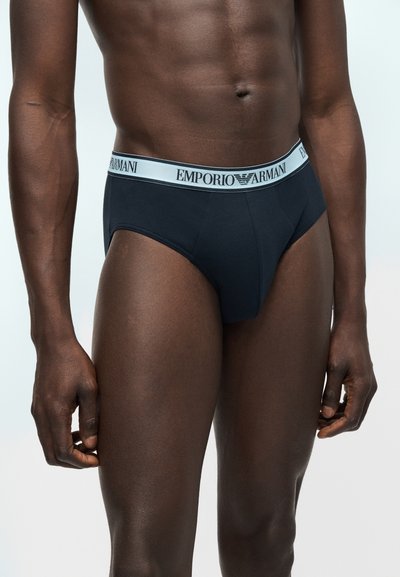 Άνδρας με κορμί που φοράει μαύρα εσώρουχα Emporio Armani με λευκή μέση και λογότυπο, στέκεται μπροστά σε ένα ανοιχτό φόντο.