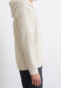 Felpa beige con tasca a marsupio e cappuccio con cordino, caratterizzata da una texture morbida e polsini a costine, indossata sopra jeans scuri.