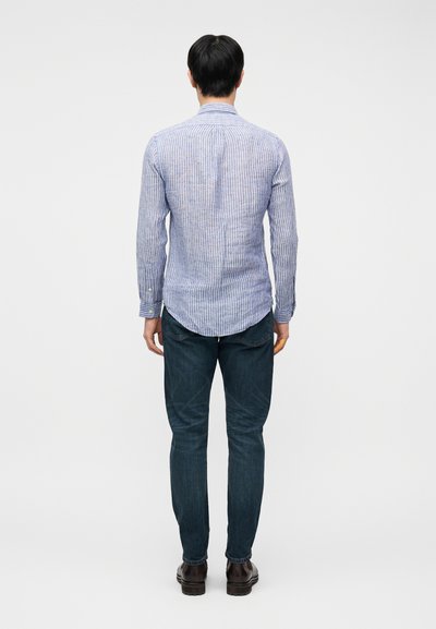 Άνδρας που φοράει σκούρο μπλε τζιν, καφέ παπούτσια και ένα ανοιχτό μπλε και άσπρο πουκάμισο με μακριά μανίκια και ρίγες, στέκεται με την πλάτη γυρισμένη σε λευκό φόντο.