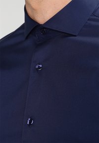 Hemd in Marineblau mit klassischem Kragen, zwei Knopfverschlüssen, glatter Textur und dezentem Glanz. Taillierte Passform betont die Silhouette.