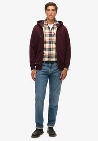 Un sweat à capuche bordeaux avec une fermeture éclair à l'avant, superposé à une chemise en flanelle à carreaux dans des tons crème, rouge et bleu, associé à un jean en denim bleu et des chaussures noires.