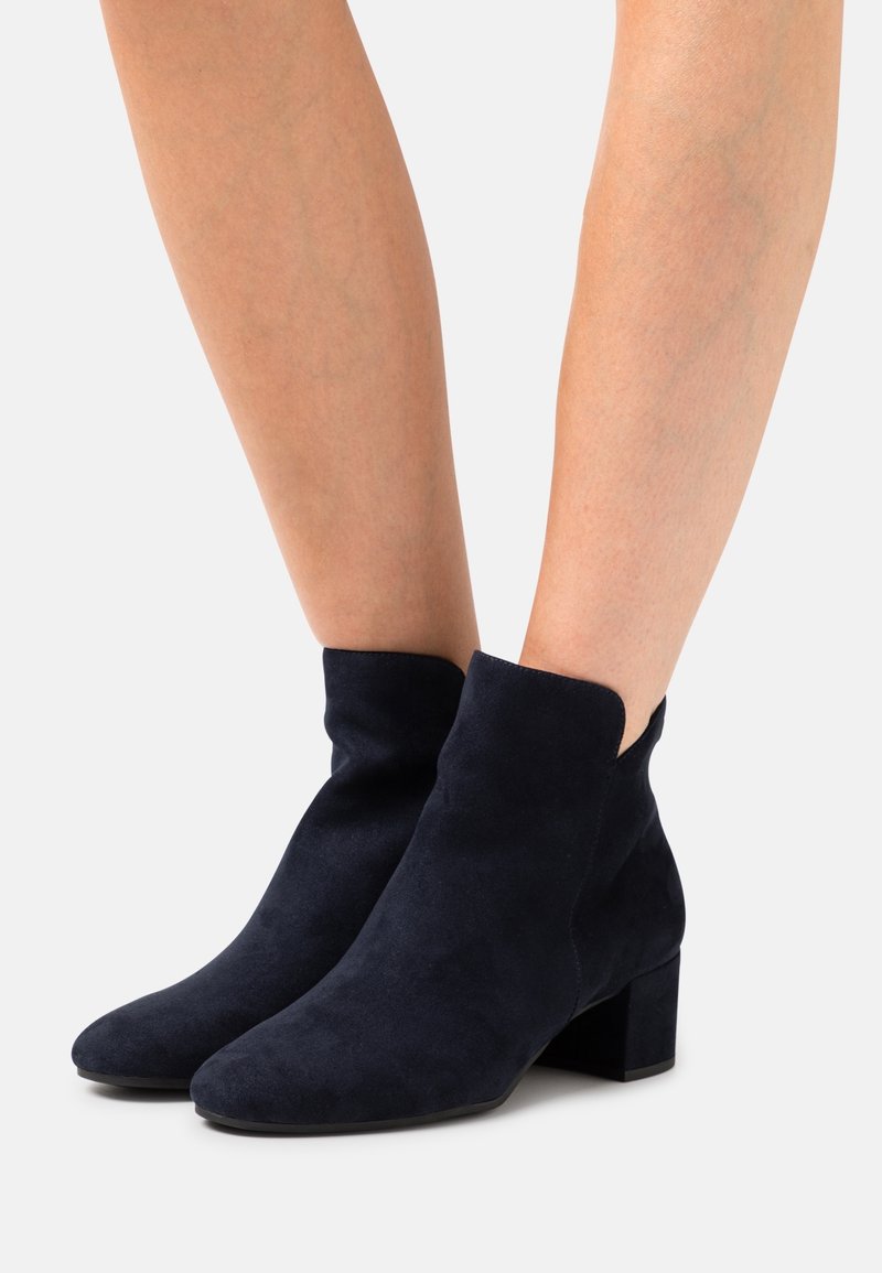 Marineblaue Wildleder-Stiefeletten mit einer schlanken, abgerundeten Spitze und einem kurzen Blockabsatz. Auffällige, geschwungene Öffnung an der Oberseite verleiht dem Schuh Details.