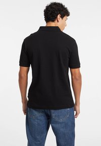 Maglietta polo nera vista da dietro, con maniche corte e colletto classico. Abbinata a jeans denim blu, che mostrano una vestibilità casual.