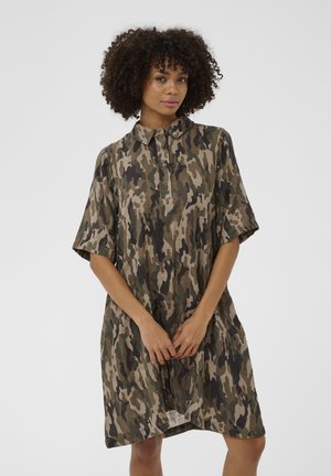 Mujer con cabello rizado que lleva un vestido de manga corta con patrón de camuflaje y cuello, de pie con las manos entrelazadas al frente contra un fondo liso.