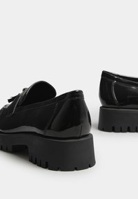 Scarpe in vernice nera con una suola spessa nera, caratterizzate da una punta arrotondata e un dettaglio a nappina decorativo sulla parte superiore.