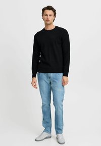 Zwarte gebreide trui met ronde hals, gecombineerd met lichtblauwe jeans en grijze sneakers. Model staat tegen een witte achtergrond.