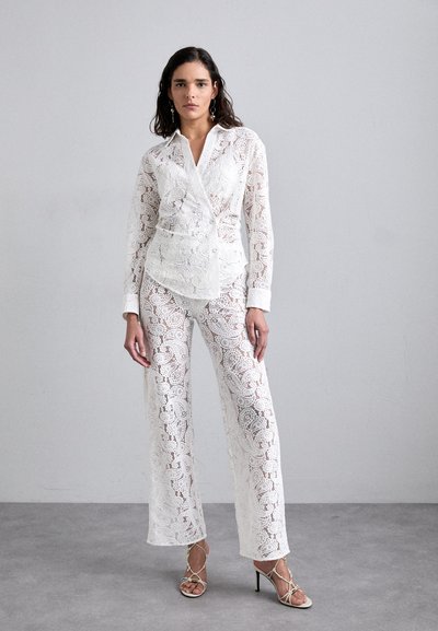 Blouse en dentelle blanche à manches longues et col, assortie à un pantalon large en dentelle. Les deux présentent des motifs floraux complexes et une texture transparente.