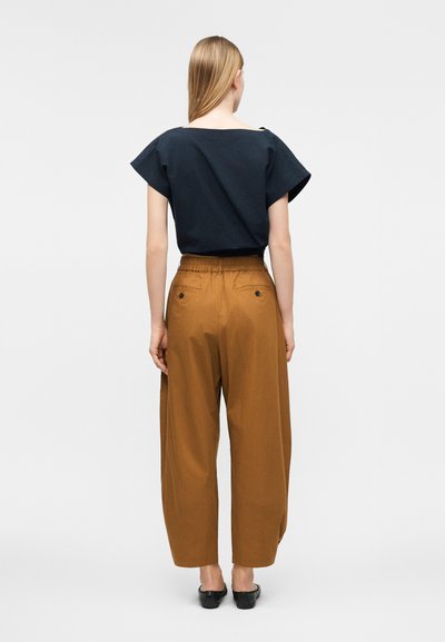 Γυναίκα με ίσια ξανθά μαλλιά φοράει μπλε σκούρο κοντομάνικο τοπ και καφέ φαρδιά παντελόνια με ψηλή μέση, στέκεται γυρισμένη προς τα πίσω με φόντο λευκό.