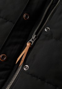 Close-up van een zwarte gewatteerde jas met een metalen ritssluiting met een bruine leren trekker en bruine drukknoopsluitingen.