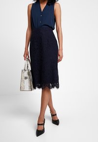 Blusa senza maniche navy con bottoni, abbinata a una gonna in pizzo navy di lunghezza al ginocchio. Gli accessori includono una borsa grigia testurizzata e tacchi neri.