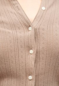 Κοντινό πλάνο μπεζ πλεκτής ζακέτας με μικρά οπτικά σχέδια και τέσσερα λευκά κουμπιά κουμπωμένα πάνω στο στήθος ενός ατόμου.