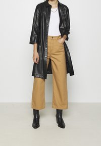 Długi czarny płaszcz z sztucznej skóry, biała koszulka i beżowe spodnie o szerokich nogawkach z przednią kieszenią. Całość uzupełniają czarne botki za kostkę.