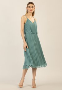 Faltenkleid in Türkis mit V-Ausschnitt, verstellbaren Trägern und mittellangem Saum. Leichter Stoff und sanfter Fall fördern die Bewegungsfreiheit.