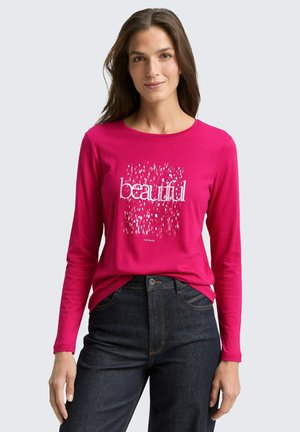 Kobieta ubrana w jaskraworóżową bluzkę z długim rękawem z napisem „beautiful” i abstrakcyjnym białym wzorem, połączoną z ciemnymi, wysokim stanem jeansami.