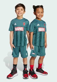 Due bambini indossano magliette da calcio turchesi con i loghi di Spider-Man e Marvel, caratterizzate da un design testurizzato. Hanno scarpe rosse e nere con calze a motivi.