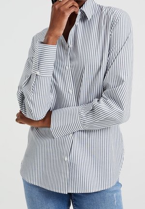 Person iført en langærmet skjorte med lodrette grå og hvide striber og blå jeans, hånden hvilende nær ansigtet og armen krydset.