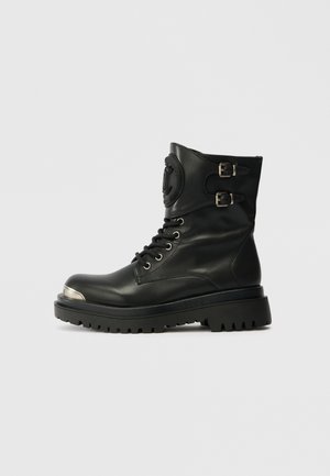 Schwarze Lederstiefelette mit metallic-farbenem Zehenkappe, klobiger Sohle, Schnürung vorne und zwei dekorativen Schnallen an der oberen Seite.