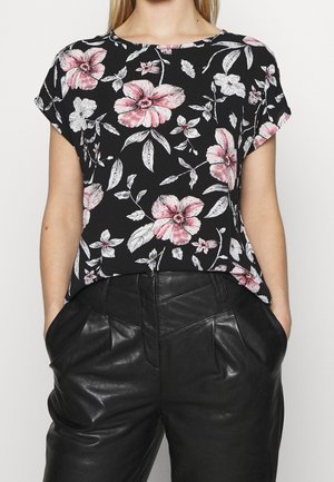 Donna che indossa un top nero a maniche corte con stampa floreale rosa e bianca, abbinato a pantaloni in pelle neri a vita alta con tasche.