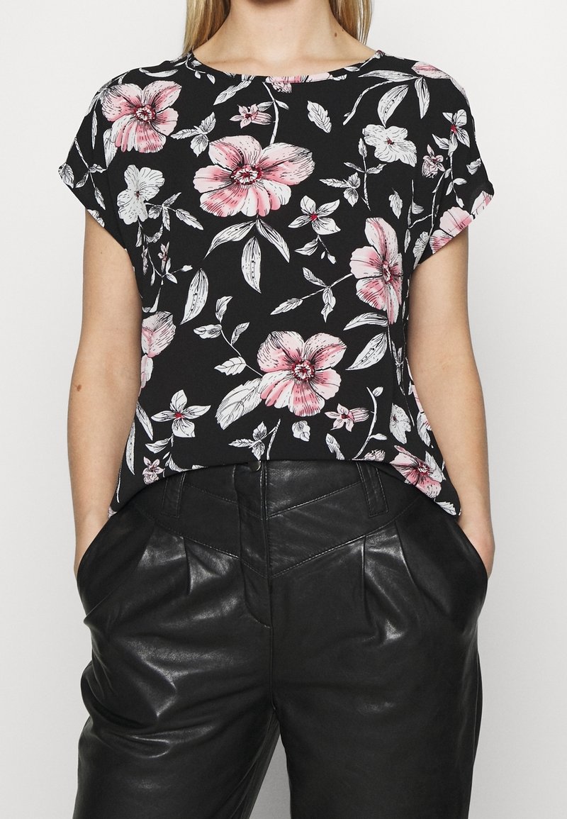 Femme portant un haut noir à manches courtes avec imprimé floral rose et blanc, associé à un pantalon en cuir noir taille haute avec poches.