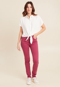 Witte korte mouwen knoopshirt, geknoopt in de taille, gecombineerd met strakke bordeauxrode broek en witte sneakers, tegen een neutrale achtergrond.