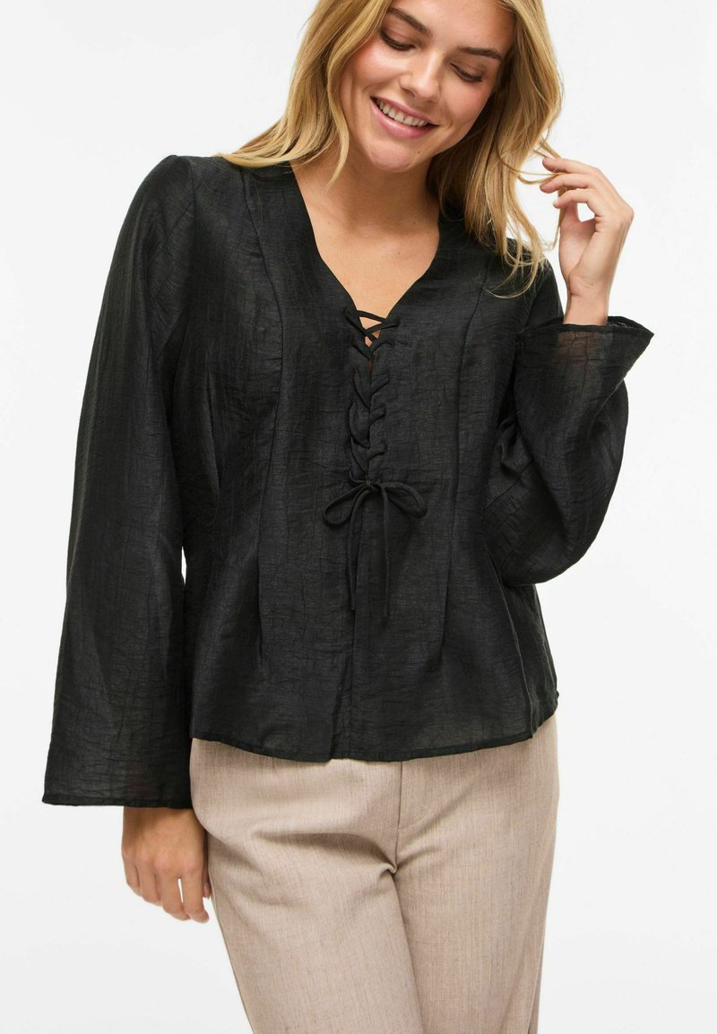 Svart bluse med knyting foran, V-hals og lange ermer. Laget av teksturert stoff. Kombinert med lyse beige bukser.