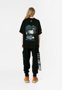 Una camiseta negra de gran tamaño presenta un gráfico de dos personajes de dibujos animados abrazándose en la parte posterior. Combinada con pantalones cargo negros y botas marrones.