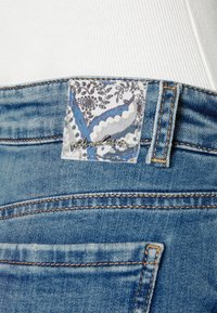 Pantalones vaqueros de denim azul con un bolsillo trasero que tiene una etiqueta de tela floral y un acento metálico plateado "Miss Sixty" en la cintura, combinados con una camiseta blanca de canalé.