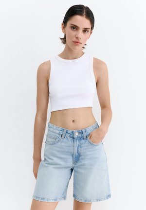 Giovane donna con capelli corti e scuri che indossa un top corto bianco senza maniche e pantaloncini di denim azzurri, con una mano in tasca.