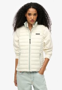 Wattierte Weste in Creme mit gesteppten Paneelen, Reißverschluss, Stehkragen und seitlichen Taschen. Mit Branding auf der linken Brust und kontrastierendem Saum.