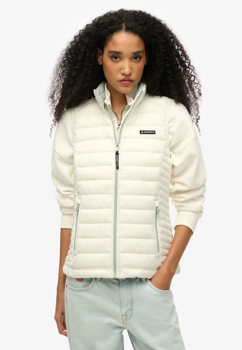 Wattierte Weste in Creme mit gesteppten Paneelen, Reißverschluss, Stehkragen und seitlichen Taschen. Mit Branding auf der linken Brust und kontrastierendem Saum.