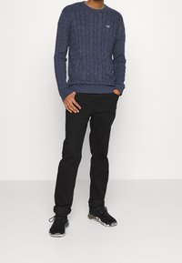 Pull en tricot torsadé bleu marine, pantalon noir et baskets noires avec une semelle à motifs. Le pull possède un col rond et des poignets côtelés.