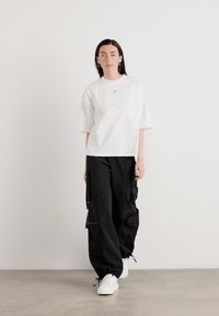 Hvit t-skjorte med korte ermer og en subtil logo, sammen med løstsittende sorte cargo-bukser og hvite sneakers på en nøytral bakgrunn.