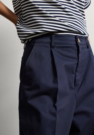 Pantaloni blu navy con pieghe frontali, tasche laterali e chiusura con bottone metallico. Abbinati a una camicia a righe blu e bianche.