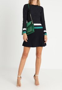 Robe noire avec un haut ajusté à motifs et une jupe évasée ; associée à un sac à main vert orné de clous ; talons à brides transparentes.