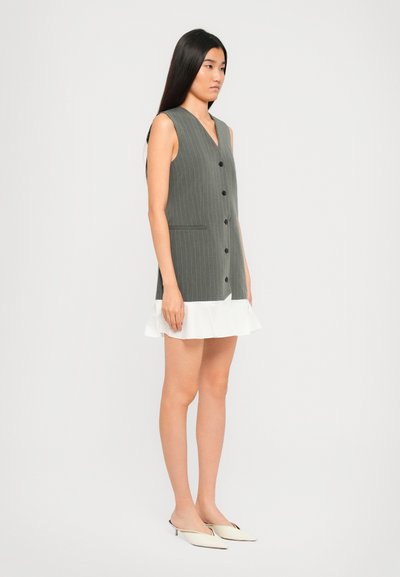 Donna in piedi con un vestito senza maniche grigio a righe con bottoni neri e orlo bianco arricciato, che indossa mule bianche con tacco basso.
