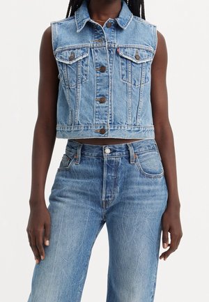 Femme portant un gilet en jean bleu clair sans manches avec fermeture à boutons et un jean taille haute assorti, sur fond blanc.