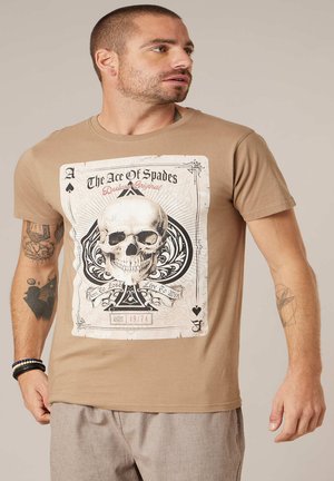Homme aux cheveux courts portant un t-shirt marron avec un motif de carte As de Pique orné d'un grand crâne et aux bras tatoués.