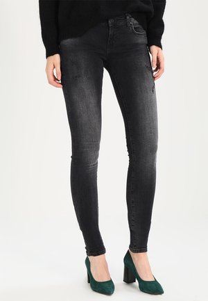 Personne portant un jean skinny noir délavé et des chaussures à talons hauts en daim vert, debout devant un fond uni.
