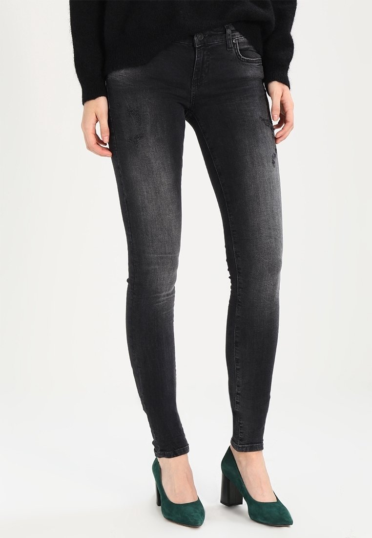 Personne portant un jean skinny noir délavé et des chaussures à talons hauts en daim vert, debout devant un fond uni.