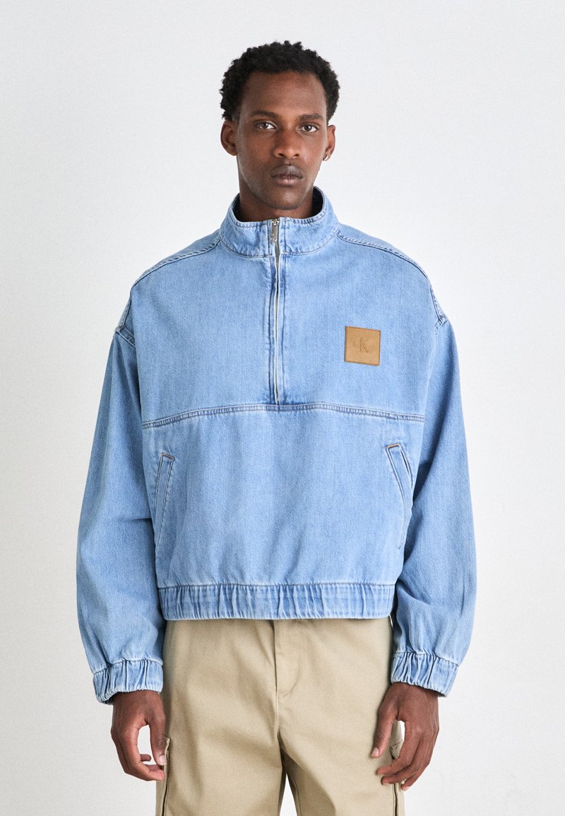 Giacca di jeans blu chiaro con colletto alto, chiusura a mezza zip sul davanti, polsini e vita elastici; presenta una toppa in pelle beige sul petto.