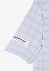Ljusblå kortärmad skjorta med rutmönster och en vit etikett med "LACOSTE" i svart text på ärmen. Strukturerad tygfinish.