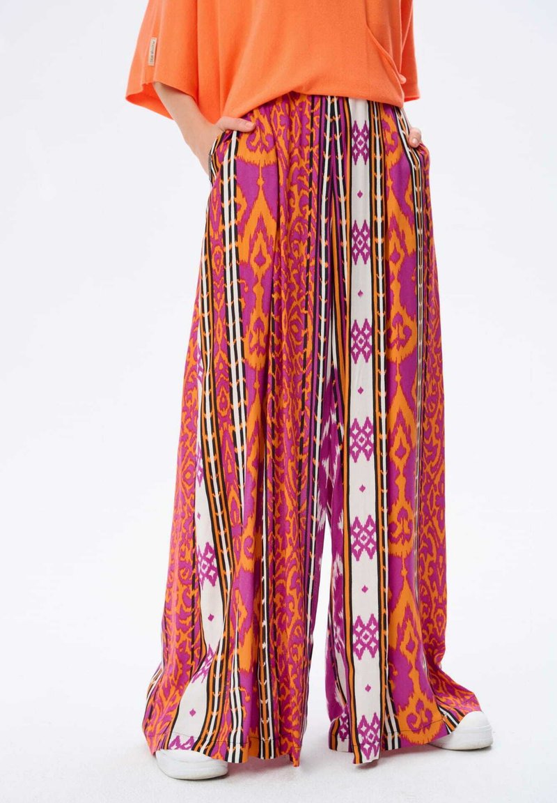 Kolorowe szerokie spodnie z wzorowanym designem w odcieniach różu, pomarańczy i bieli z czarnymi akcentami. Materiał wydaje się lekki i przewiewny.