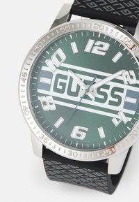 Silberne Uhr mit grünem Ziffernblatt, großen weißen Ziffern und "GUESS"-Logo. Schwarzes Gummiband mit geometrischem Muster. Rundes Gehäuse aus Edelstahl.