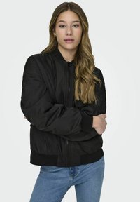 Juodas bomberio tipo švarkas su užtrauktuku priekyje, susiraukšlėjusiomis rankovėmis ir apačioje. Yra glotnus, blizgus tekstūrinis paviršius ir laisvas siluetas. Suderinta su mėlynomis džinsomis.